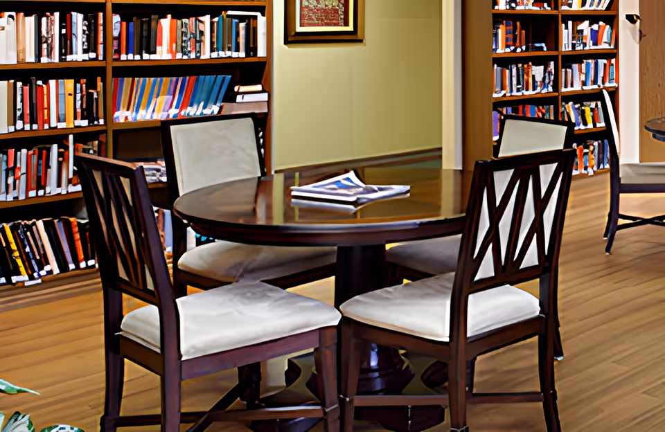 A cozy library or reading area with a round wooden table surrounded by four cushioned chairs. Bookshelves filled with books line the walls, and a framed picture hangs on the wall above the table. The floor is wooden, and a folded newspaper rests on the table.