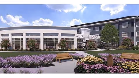 Outdoor garden area at Watercrest Richmond featuring a curved building with large windows, landscaped flower beds with purple and yellow flowers, green shrubs, trees, and wooden benches along a paved walkway under a partly cloudy sky.