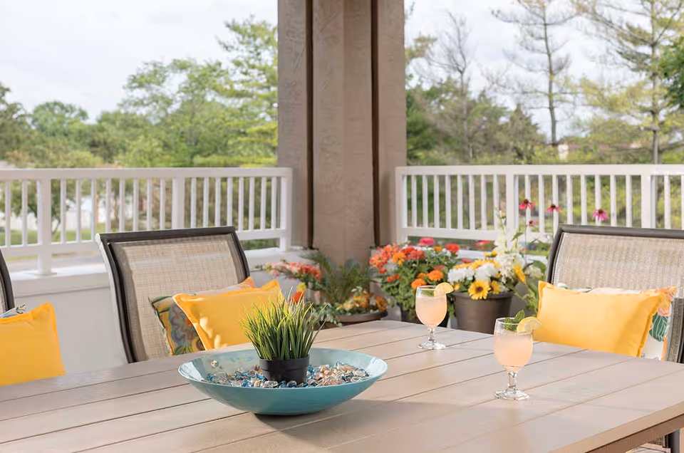 Outdoor patio area with a table and chairs featuring yellow cushions. On the table, there is a decorative bowl with a small potted plant and two glasses of a pink beverage garnished with lemon and lime slices. The background shows a white railing and various colorful flowers with green trees beyond.