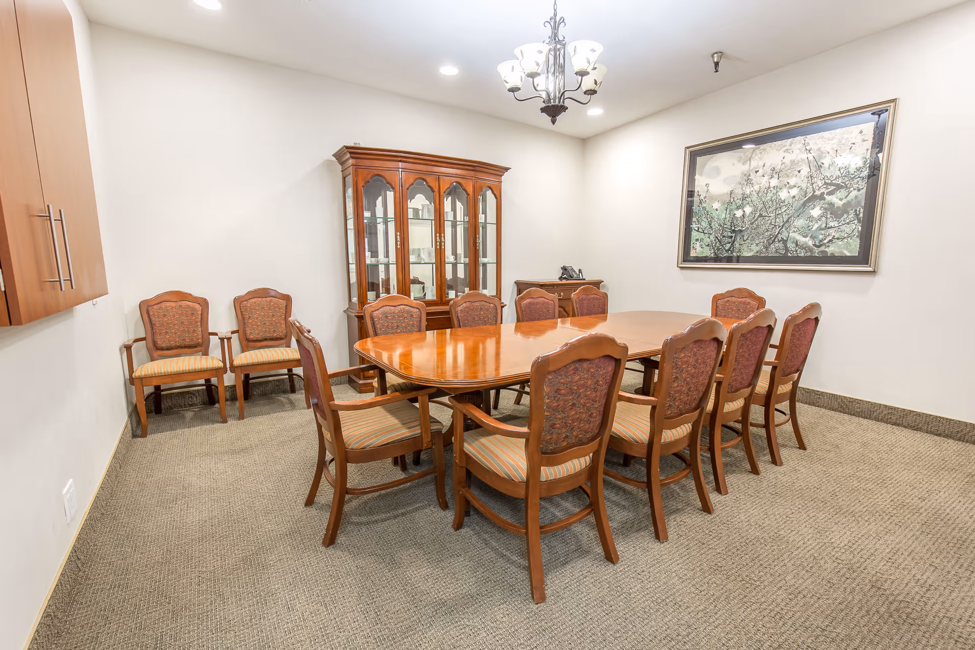 A well-lit dining room with a large wooden dining table surrounded by ten upholstered chairs. There is a wooden china cabinet with glass doors against the back wall, a small side table with a telephone, a chandelier hanging from the ceiling, and a framed artwork on the wall. The room has beige carpet and white walls.