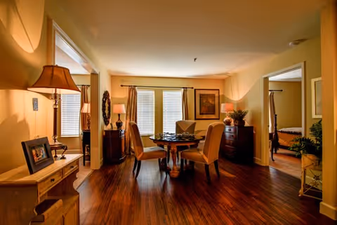 A warmly lit interior room with wooden flooring featuring a round table with four chairs in the center. The room has two windows with blinds and curtains, two lamps on side tables, framed artwork on the walls, and doorways leading to other rooms including a bedroom visible on the right.