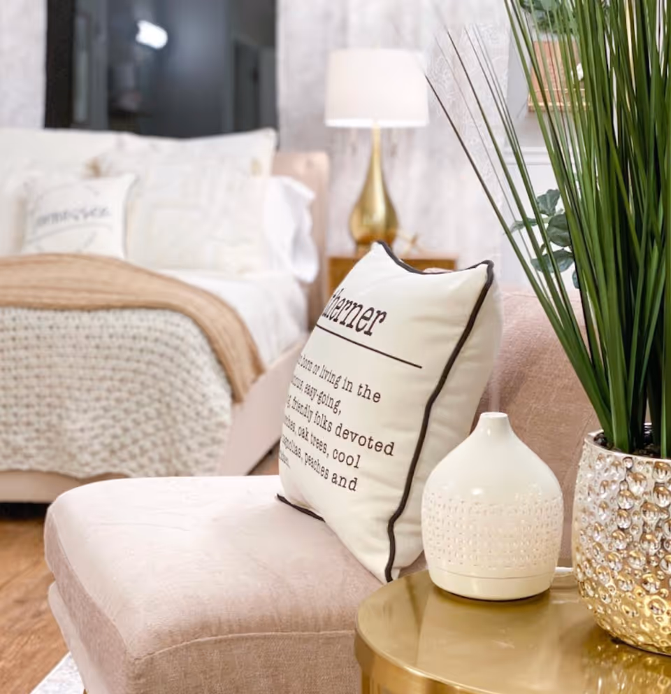 A cozy bedroom corner featuring a beige cushioned chair with a decorative pillow, a small round gold side table with a white ceramic vase and a gold textured planter holding tall green plants. In the background, a neatly made bed with white and beige bedding and pillows is visible, along with a gold lamp on a wooden nightstand.