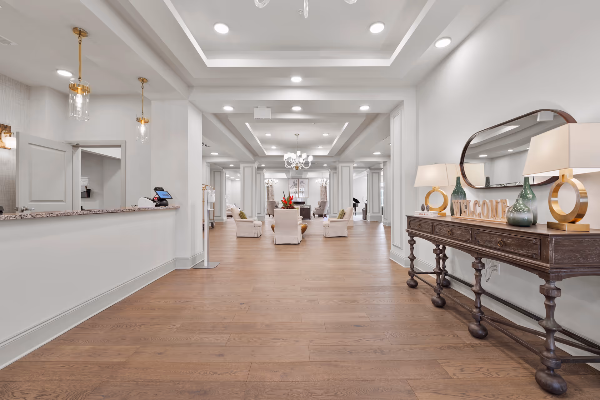 Spacious and well-lit interior of a senior living facility with wooden flooring, white walls, and a high ceiling with recessed lighting. On the right side, there is a dark wooden console table with two lamps, decorative vases, and a 'WELCOME' sign in front of a large oval mirror. On the left side, there is a reception desk with a granite countertop and pendant lights hanging above. In the background, there are several armchairs arranged around a coffee table with a flower arrangement, and a chandelier hanging from the ceiling.