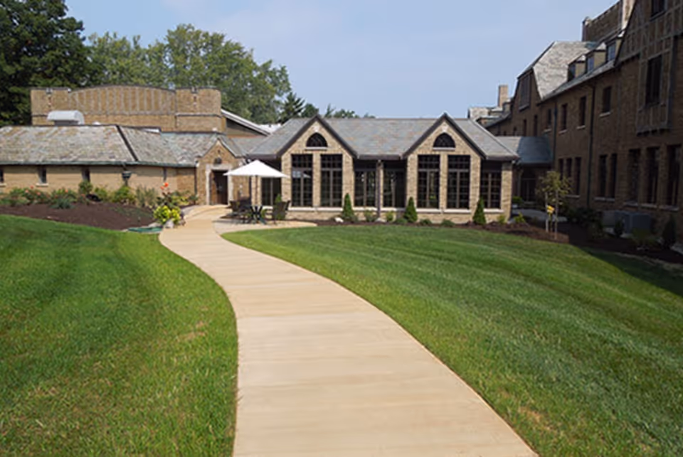A paved walkway curves through a well-maintained green lawn leading to a stone building with large windows and a slate roof. There is an outdoor seating area with a white umbrella near the entrance, and trees and shrubs surround the building under a clear sky.