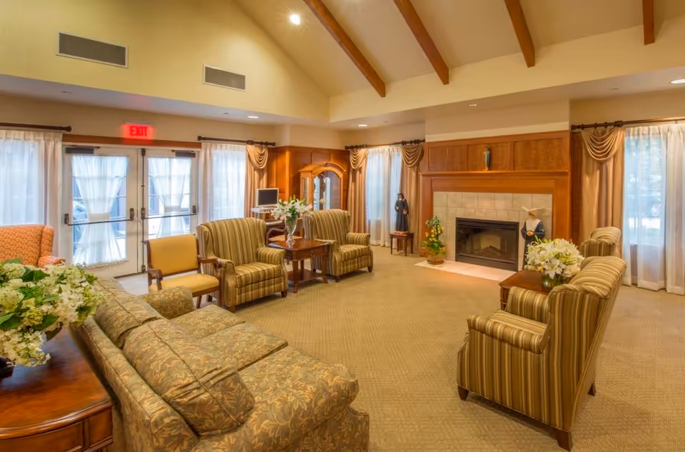 A warm, well-furnished senior living lounge with sofas and armchairs arranged around a fireplace and coffee tables.