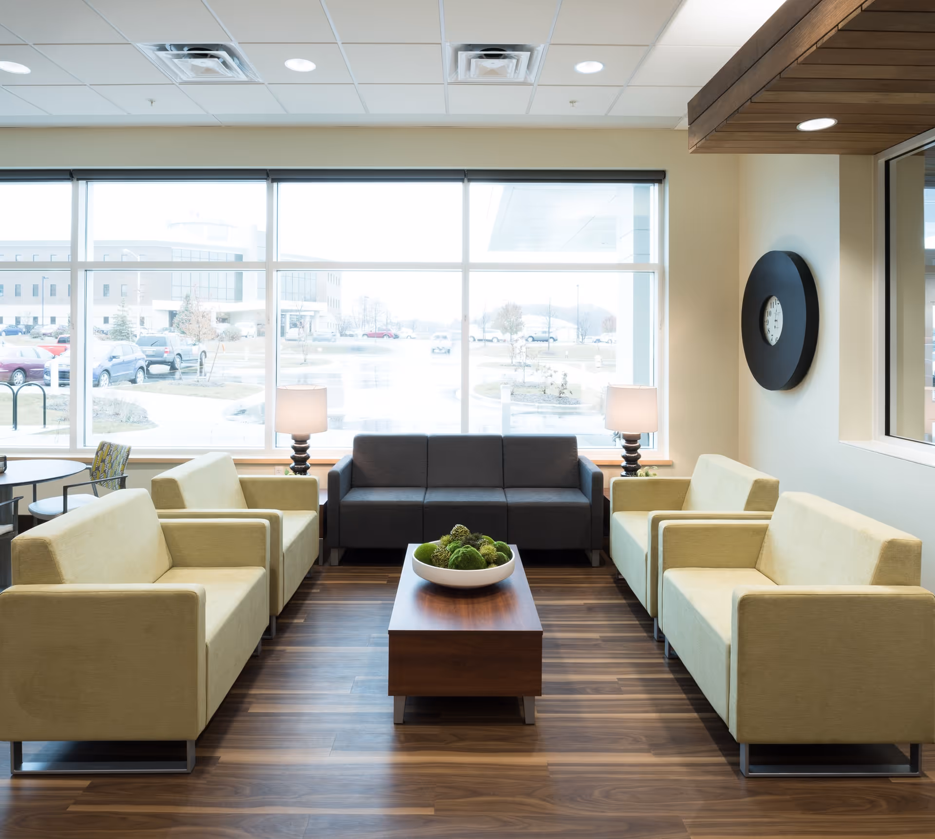 A bright and modern sitting area with four beige armchairs and one dark gray sofa arranged around a wooden coffee table with a decorative bowl of green moss balls. Two table lamps with beige shades are placed on either side of the sofa. Large windows behind the seating area provide a view of a parking lot and buildings outside. A round black wall clock is mounted on the right wall.