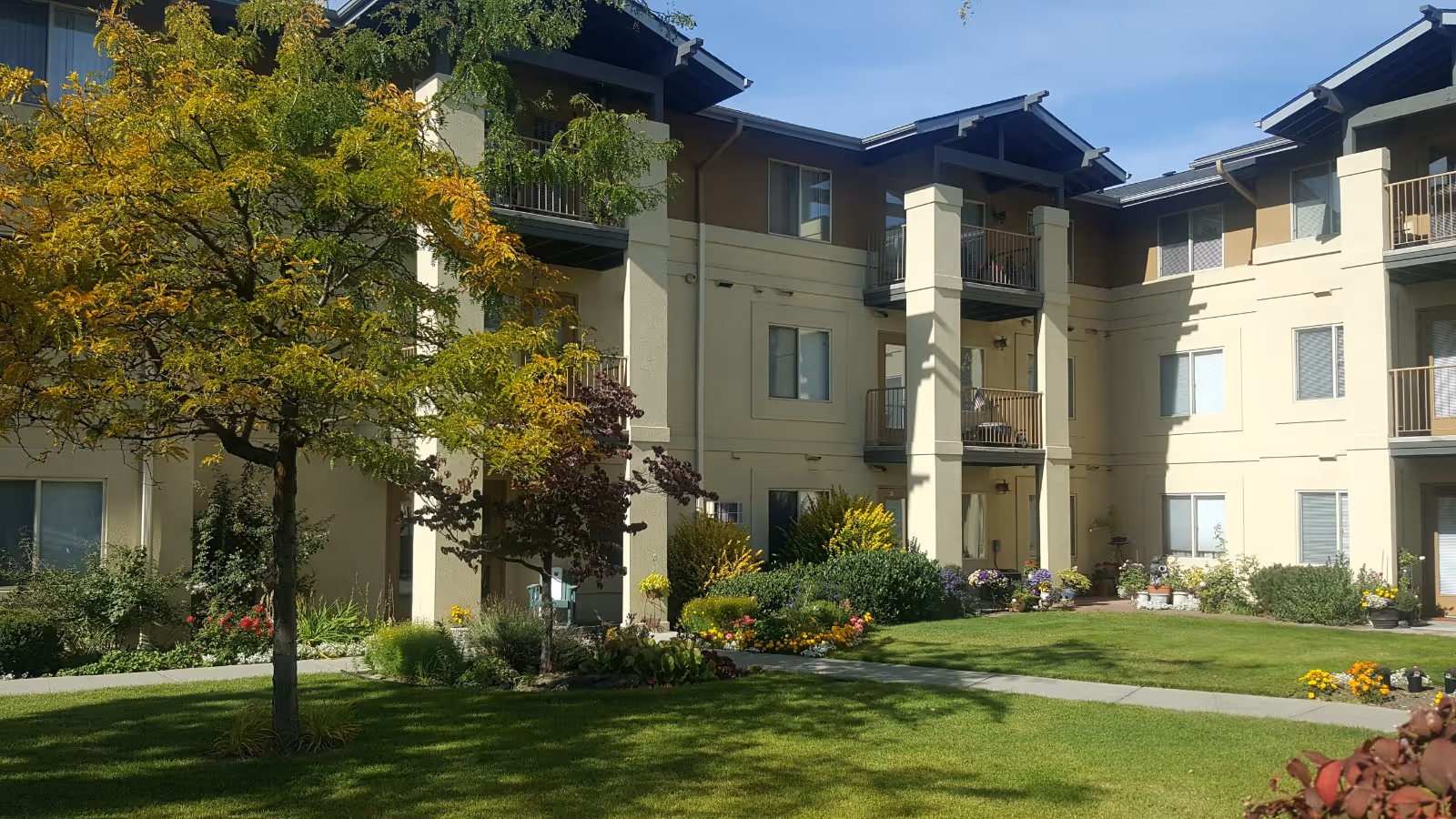 Courtyard with green lawn, trees, and flowerbeds in front of a three-story beige senior living building with balconies.