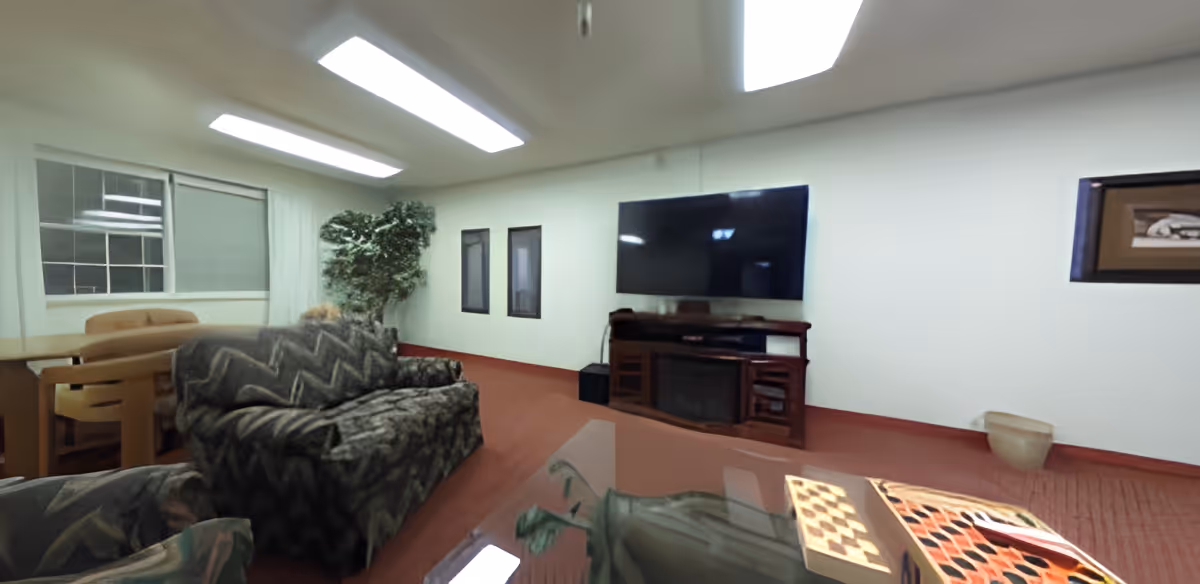 A senior living facility common area with patterned sofas, a large flat-screen TV mounted on the wall above a wooden cabinet with a fireplace, a glass coffee table with a checkerboard game, a dining table with chairs near a window, and a potted plant in the corner. The room has white walls, red carpet, and fluorescent ceiling lights.