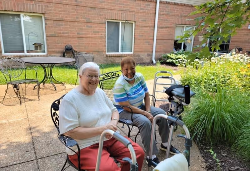 Two elderly women sitting on metal chairs in an outdoor patio area with a brick building and windows in the background. One woman is wearing a white sweater and red pants, and the other is wearing a striped shirt and gray pants. Both have walkers in front of them, and there is greenery and flowers nearby.