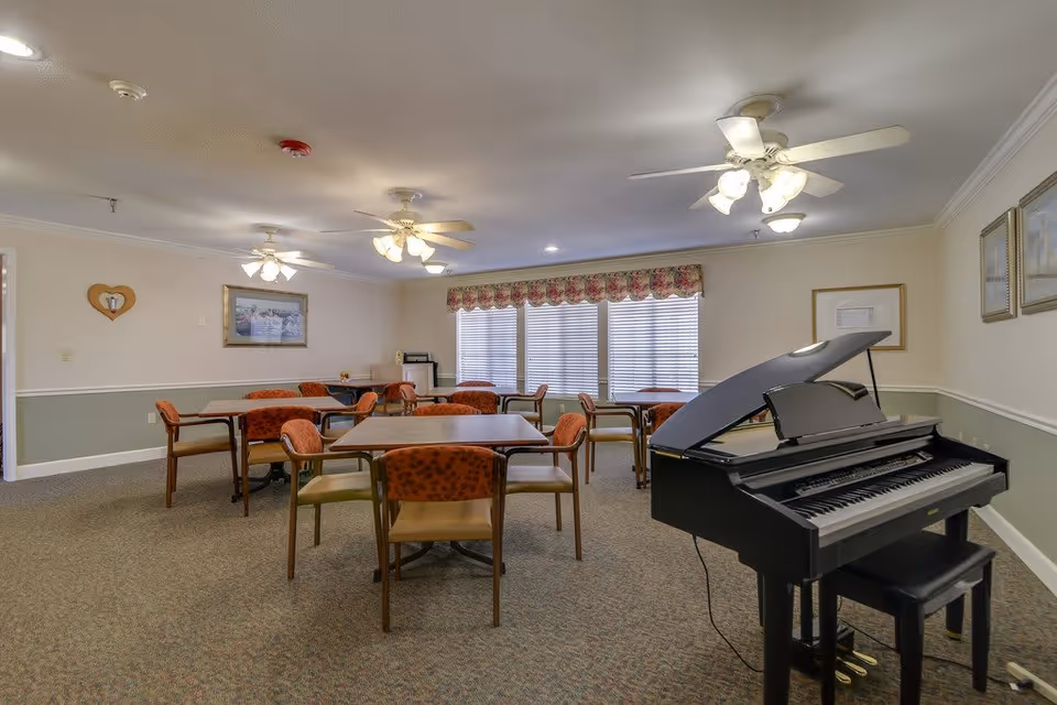 A well-lit room with several tables and chairs arranged for group seating. The room features a black grand piano with a matching bench on the right side. The walls are decorated with framed pictures and a heart-shaped wall hanging. Large windows with floral valances allow natural light to enter the space. Ceiling fans with lights are mounted on the ceiling.