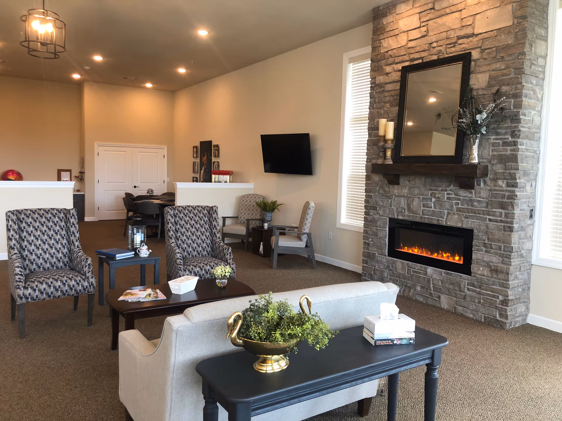 A cozy senior living facility common area with patterned armchairs, a beige sofa, a dark wooden coffee table, and a black console table with a decorative plant and tissue box. The room features a stone fireplace with a mirror and candles on the mantel, a wall-mounted TV, and large windows with blinds letting in natural light.