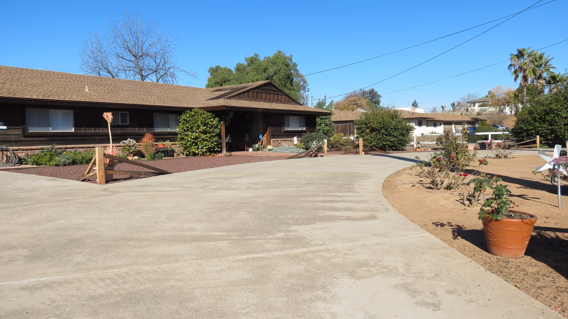 Exterior view of a single-story brown wooden building with a sloped roof, surrounded by a concrete driveway and landscaped garden areas with bushes and potted plants under a clear blue sky.