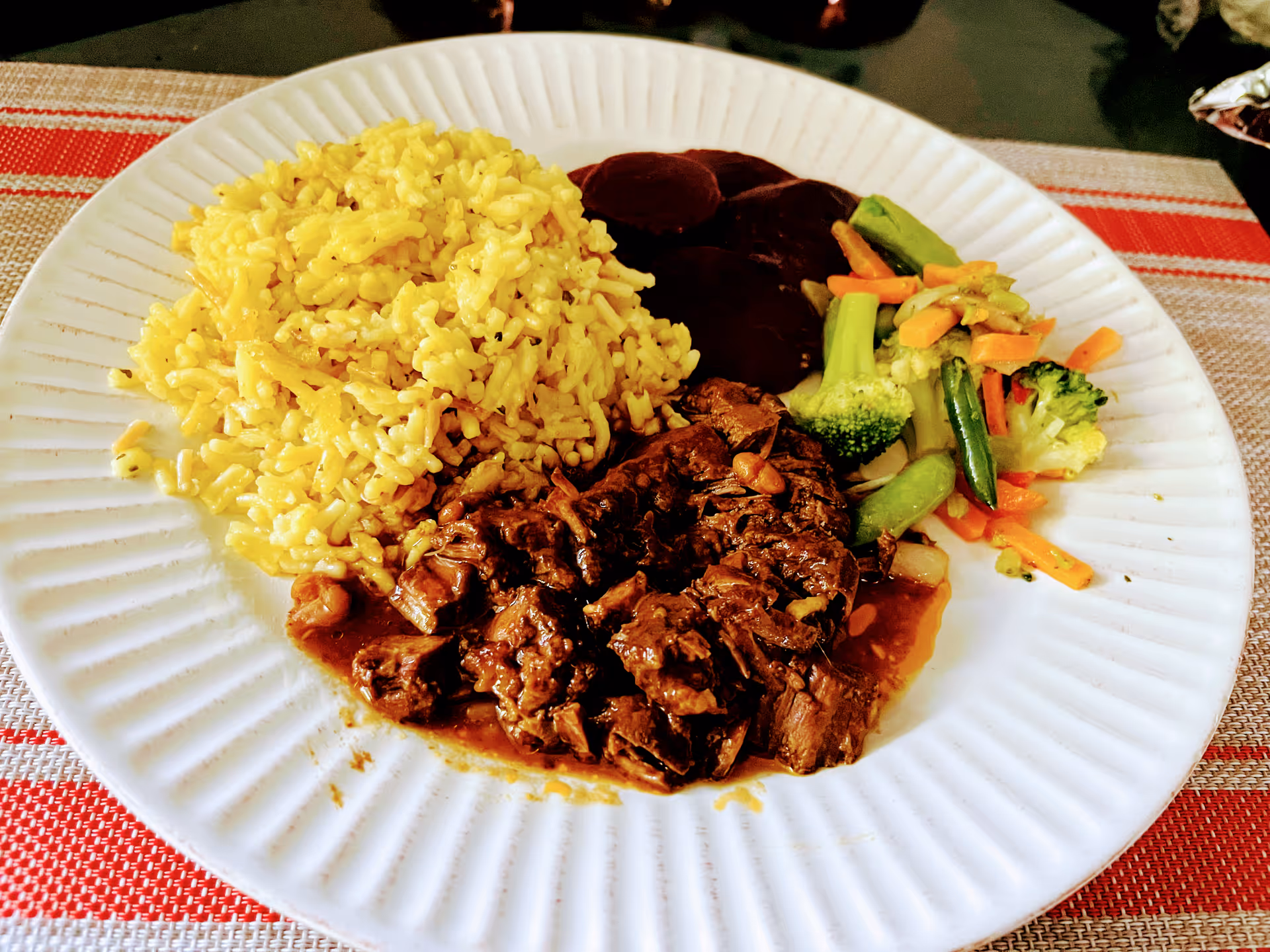A white plate with a serving of yellow rice, cooked beef in brown sauce, sliced beets, and mixed steamed vegetables including broccoli, carrots, and snap peas on a red and beige striped placemat.