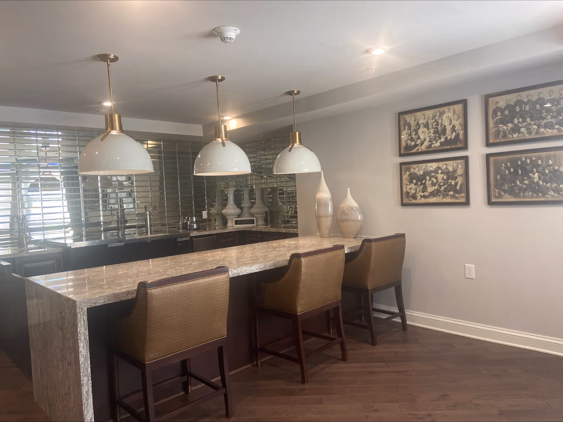 Modern kitchen area with a marble countertop island and three brown cushioned bar stools. Three large white pendant lights hang above the island. The back wall features a reflective tile backsplash and a sink. Decorative vases are placed on the countertop near the wall, which is adorned with four framed vintage black and white group photos.
