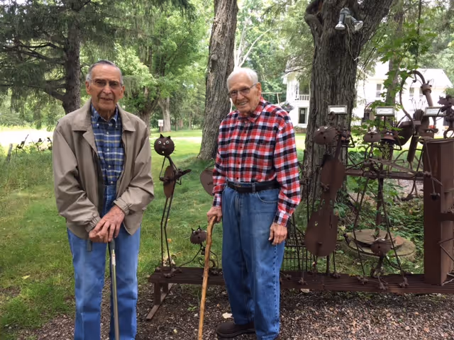 Two elderly men standing outdoors on a grassy area with trees and metal sculptures shaped like musicians in the background. Both men are using walking sticks and smiling at the camera.