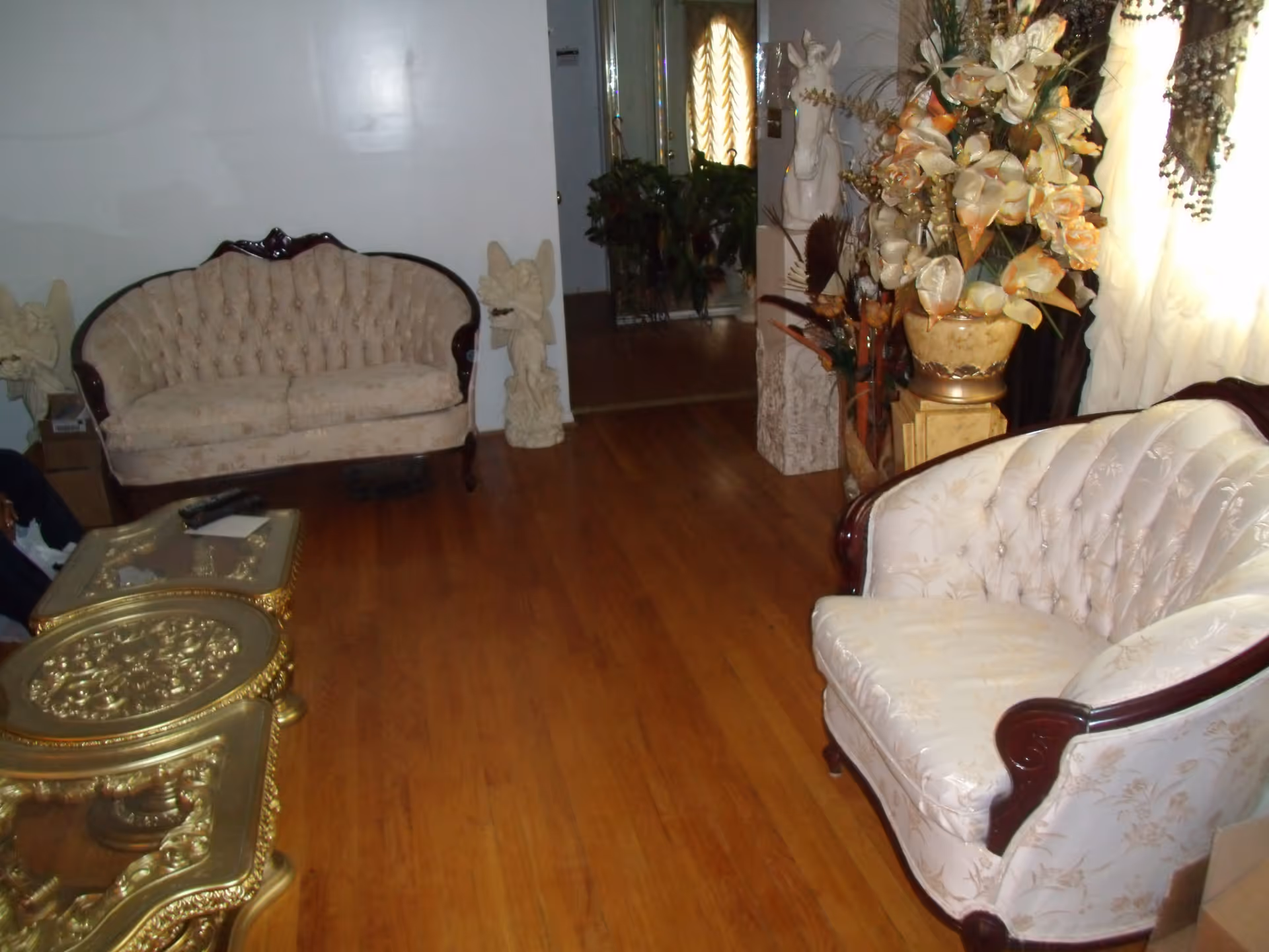 A living room with two vintage-style cream upholstered sofas with dark wood trim, a gold ornate coffee table, wooden flooring, decorative statues, and a large floral arrangement near a window with sheer curtains.
