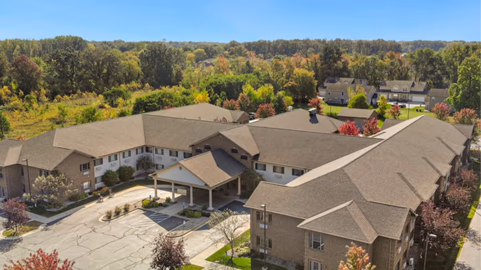 Aerial view of AVIVA Valparaiso senior living facility surrounded by trees with autumn foliage. The building has a large, multi-wing structure with a covered entrance and parking area in front. The surrounding area includes greenery and additional residential buildings in the background.
