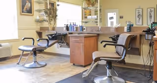 Interior of a hair salon area with two salon chairs facing mirrors mounted on the wall. The space includes a wooden cabinet with hair care products on the countertop and shelves. The room has light-colored walls, framed artwork, and a window letting in natural light.