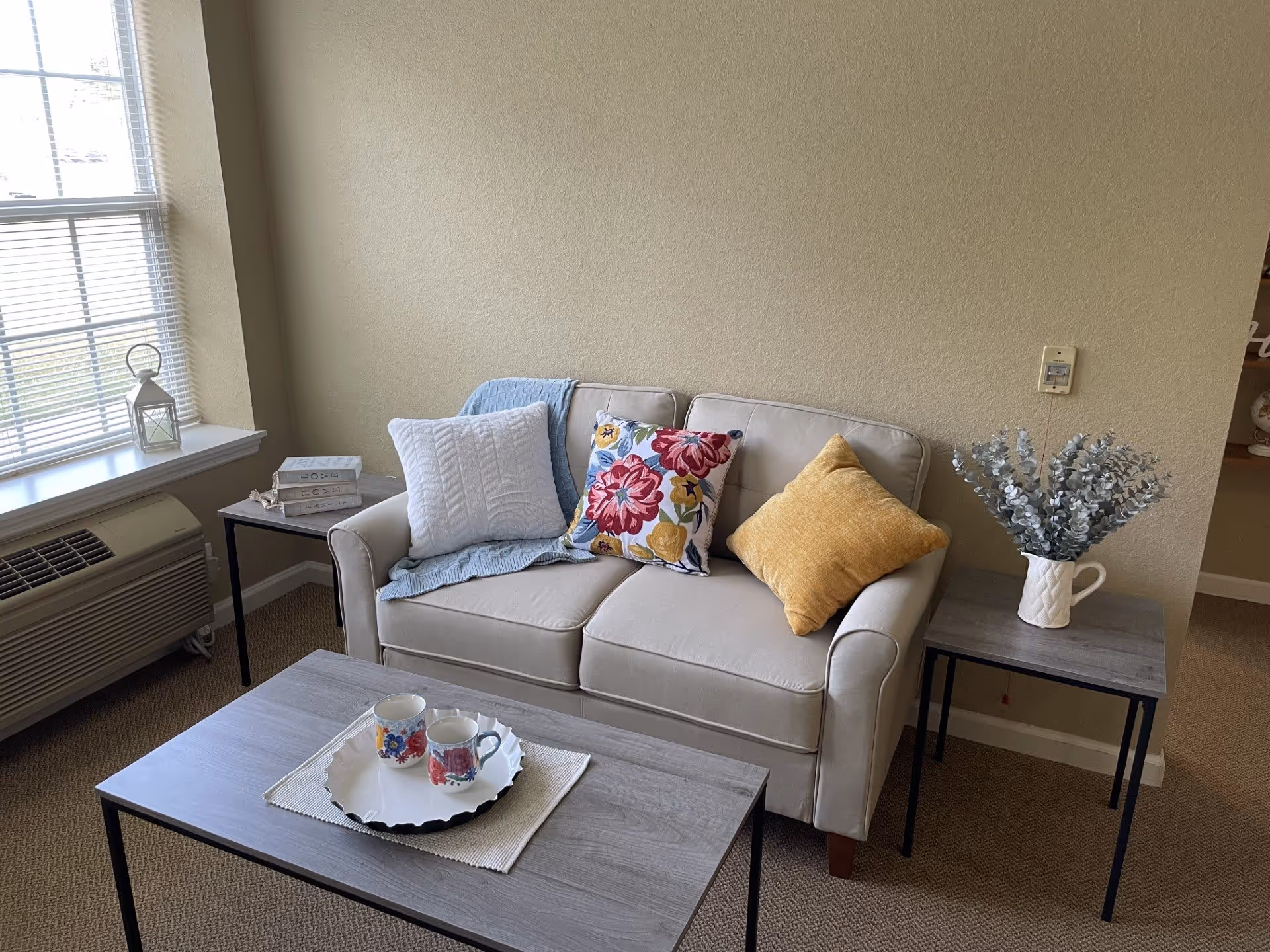 A cozy living room area with a beige loveseat adorned with three decorative pillows in white, floral, and yellow colors. A light blue throw blanket is draped over the loveseat. In front of the loveseat is a wooden coffee table with a white tray holding two floral mugs. To the right of the loveseat is a small side table with a white vase containing silver foliage. To the left is another small table with stacked books and a white lantern on the windowsill. The room has beige walls and carpet, with a window letting in natural light and an air conditioning unit below it.