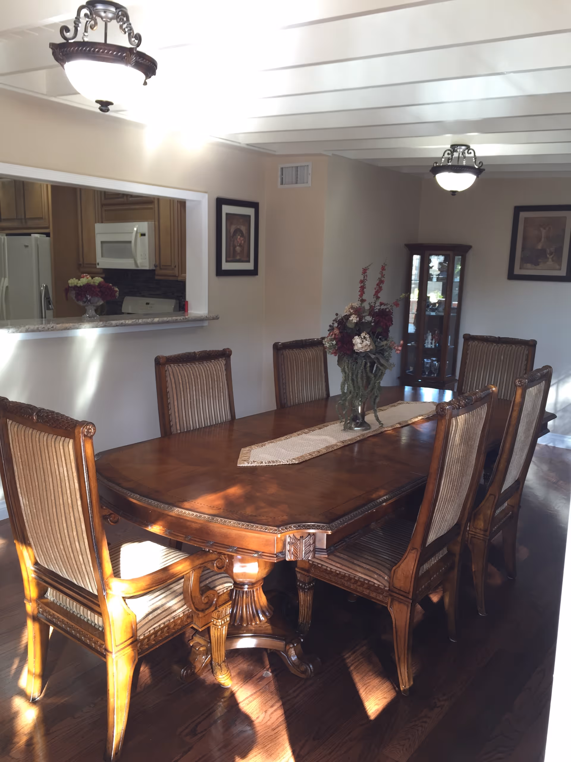 A dining room with a large wooden dining table surrounded by six upholstered chairs. The table has a decorative runner and a floral centerpiece. The room has hardwood floors, two ceiling light fixtures, and a window opening to a kitchen area with visible appliances. There are framed pictures on the walls and a glass display cabinet in the corner.