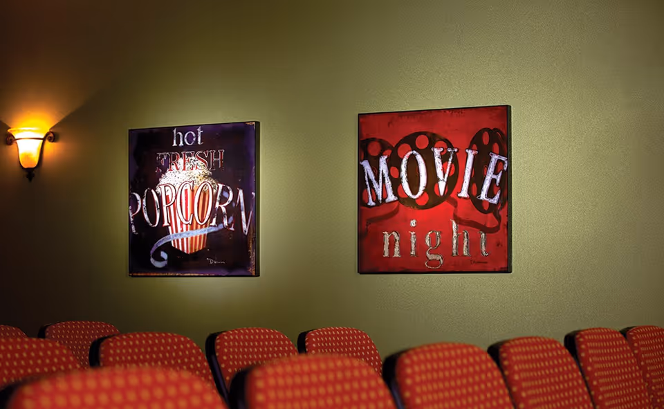 Interior view of a small movie theater or screening room with rows of red cushioned seats. Two decorative wall signs are visible on a green wall, one reads 'hot FRESH POPCORN' with an image of popcorn, and the other reads 'MOVIE night' with film reels in the background. A wall sconce light fixture is also visible on the left side.