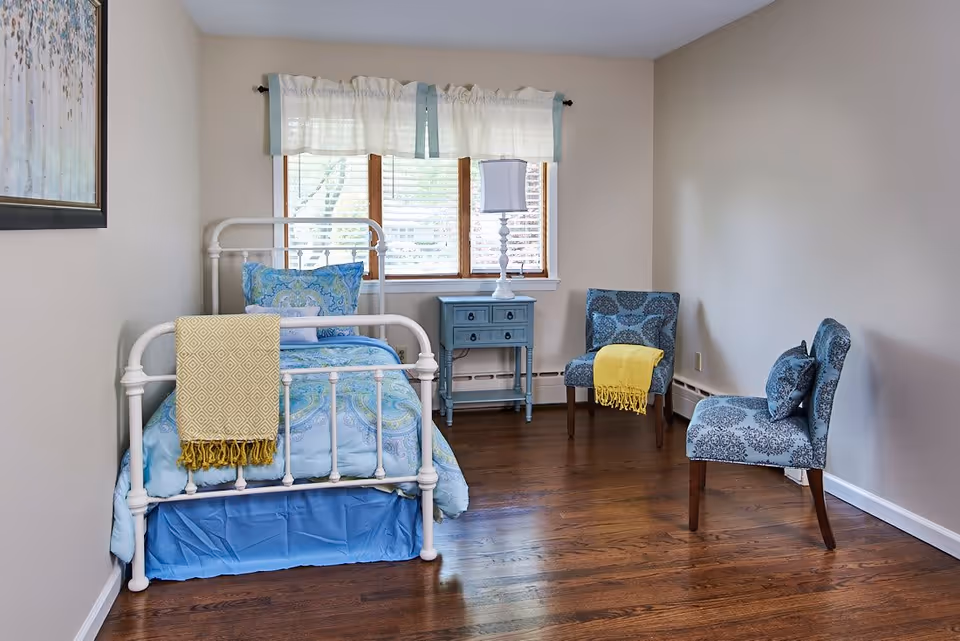A small bedroom with a white metal bed frame, blue and green patterned bedding, and a yellow throw blanket draped over the footboard. There is a blue nightstand with a white lamp in front of a window with white curtains. Two blue patterned chairs, one with a yellow throw blanket, are placed against the beige walls on a wooden floor.