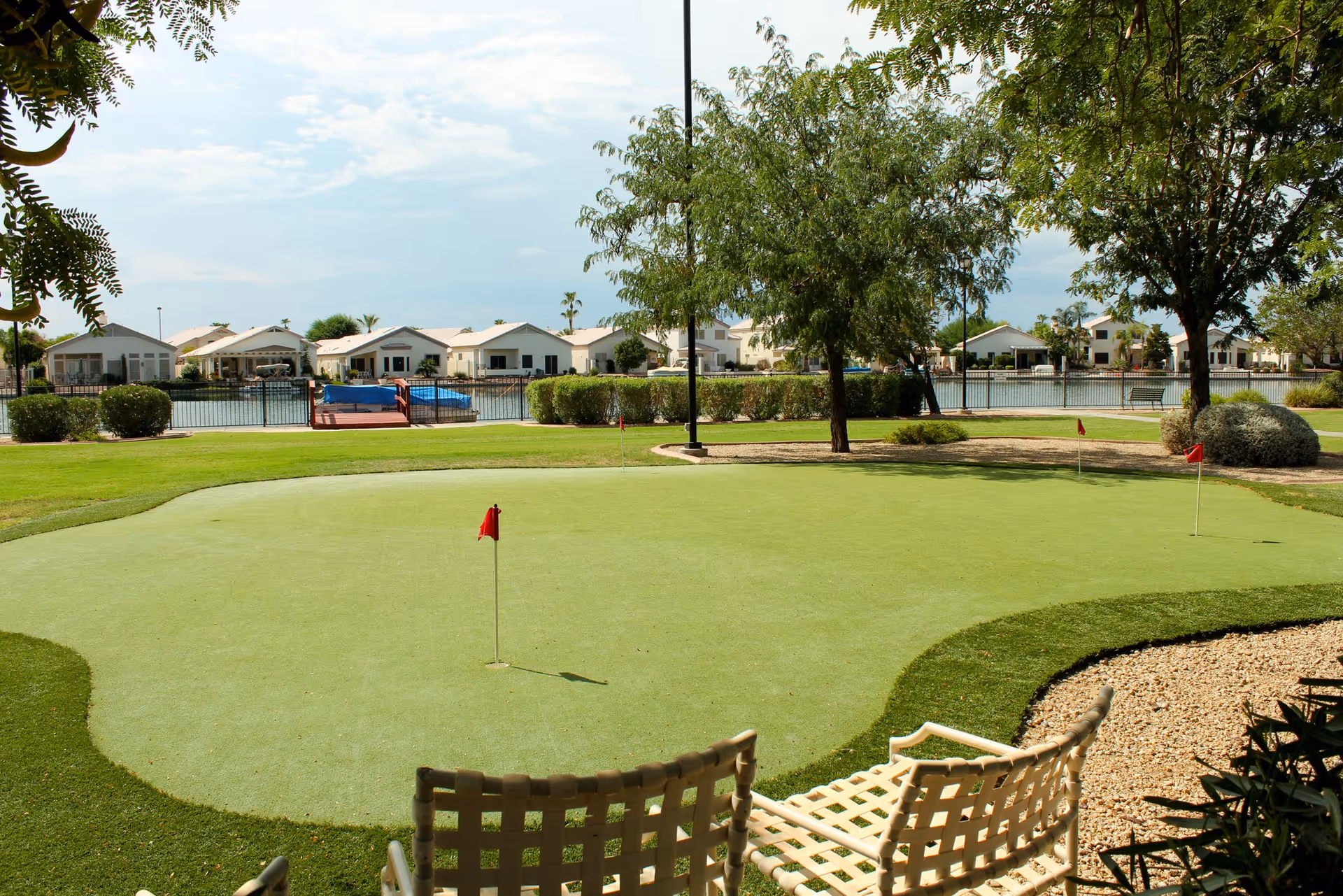 A putting green with three red flags surrounded by grass, trees, and shrubs. In the background, there is a body of water with houses along the shore under a partly cloudy sky. Two white woven chairs are visible in the foreground.