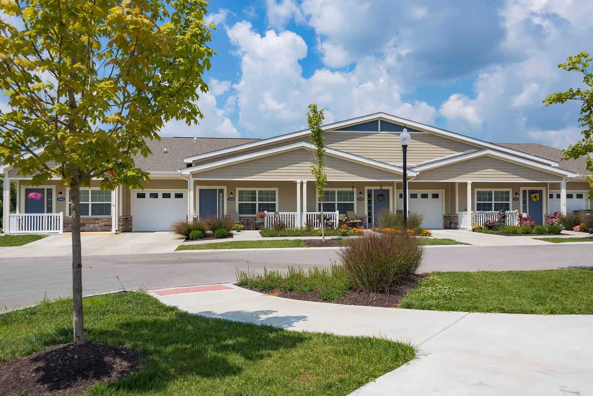 Exterior view of a single-story residential building with multiple units, each having a small porch, garage, and front door. The building is beige with white trim and surrounded by well-maintained landscaping including grass, bushes, and small trees under a partly cloudy sky.