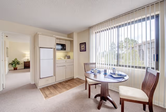 A small dining area with a round wooden table set for two with cups and napkins, two wooden chairs with cream cushions, a large window with vertical blinds letting in natural light, and a kitchenette with a refrigerator, microwave, and cabinets. A doorway leads to a bedroom with a plant and nightstand visible.