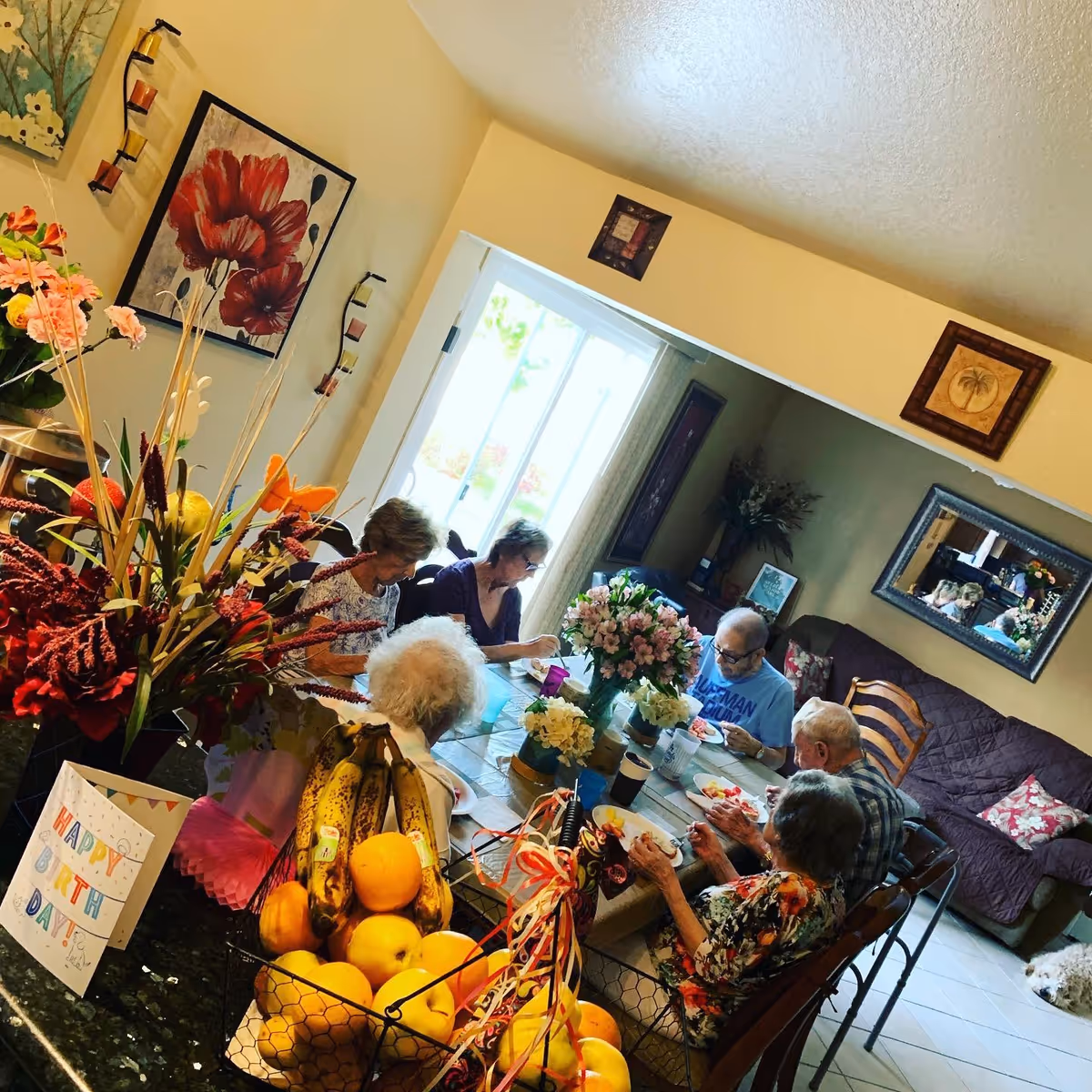 A group of elderly people sitting around a dining table in a cozy room, enjoying a meal together. The table is decorated with flowers and surrounded by chairs. In the foreground, there is a basket of bananas and oranges, and a colorful Happy Birthday card is visible. The room has wall art, a mirror, and a couch with pillows in the background.