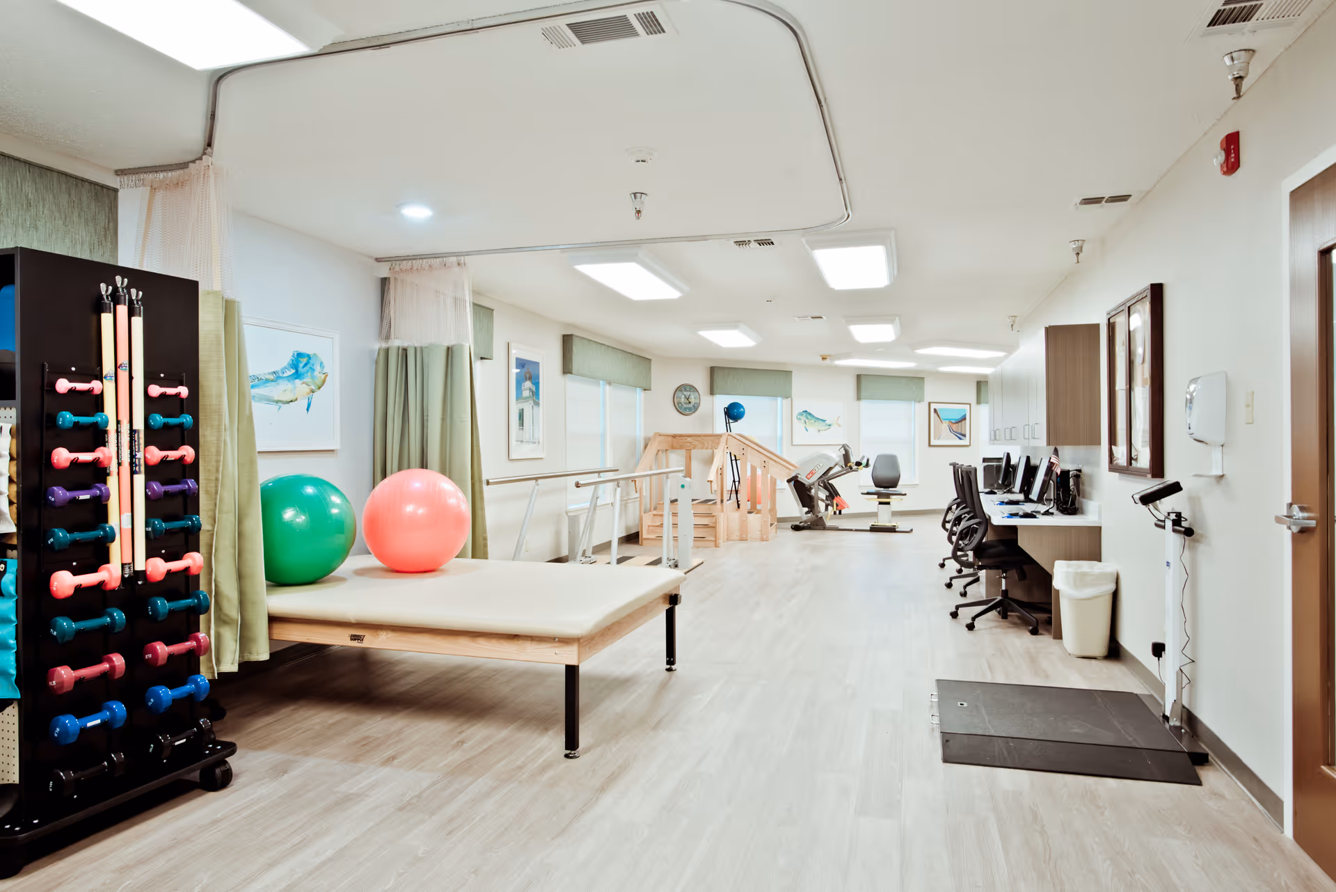 A bright and spacious physical therapy room with light wood flooring and white walls. The room contains exercise equipment including colorful dumbbells on a rack, two large exercise balls on a padded table, parallel bars for walking practice, a stationary exercise bike, and a set of stairs with handrails. There are several computer workstations with chairs along one wall, and framed artwork of birds on the walls. The ceiling has multiple rectangular fluorescent lights.