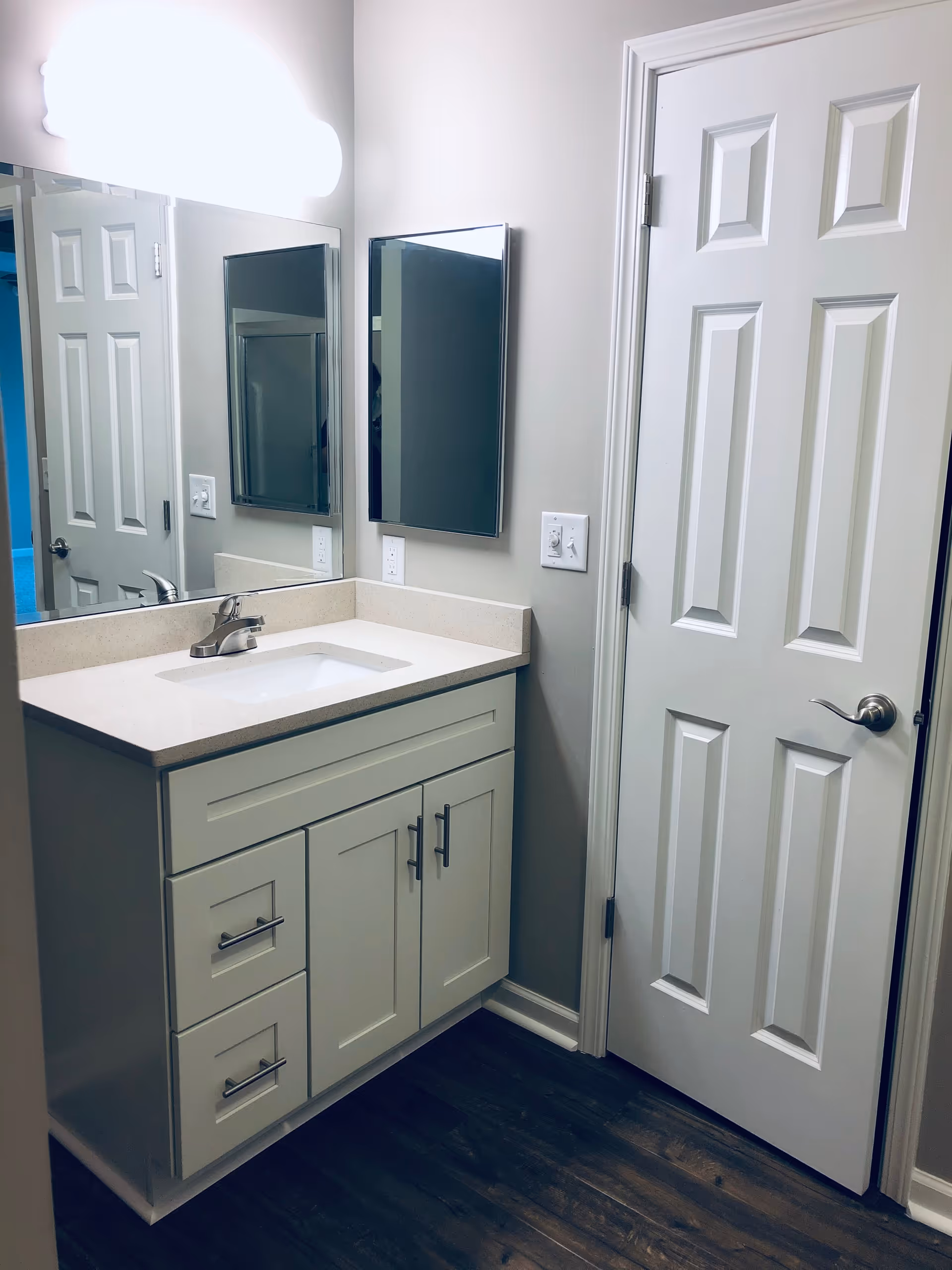 A clean and modern bathroom vanity area with a white countertop, an under-mount sink, and silver faucet. There are white cabinets and drawers below the countertop. A large mirror is mounted on the wall above the sink, with a smaller mirrored medicine cabinet to the right. A white door with a silver handle is next to the vanity. The floor is dark wood, and the walls are painted light gray.