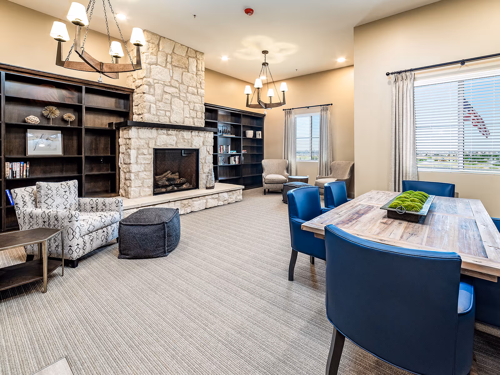 Bright communal lounge with a stone fireplace, built-in bookshelves, armchairs, and a wooden table surrounded by blue chairs.