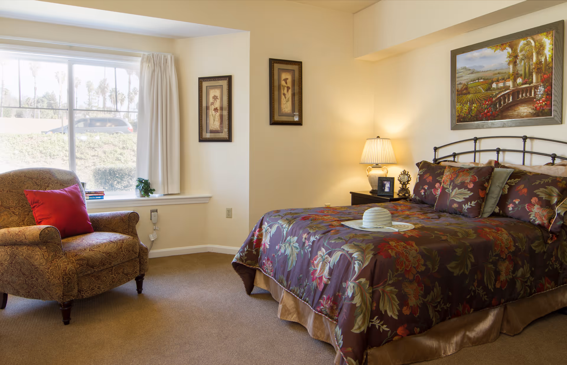 A cozy bedroom with a large bed covered in a floral patterned comforter and matching pillows. A hat rests on a tray on the bed. To the left, there is a patterned armchair with a red pillow next to a window with white curtains. The walls are decorated with framed artwork, and a bedside table with a lamp and a small clock is visible.
