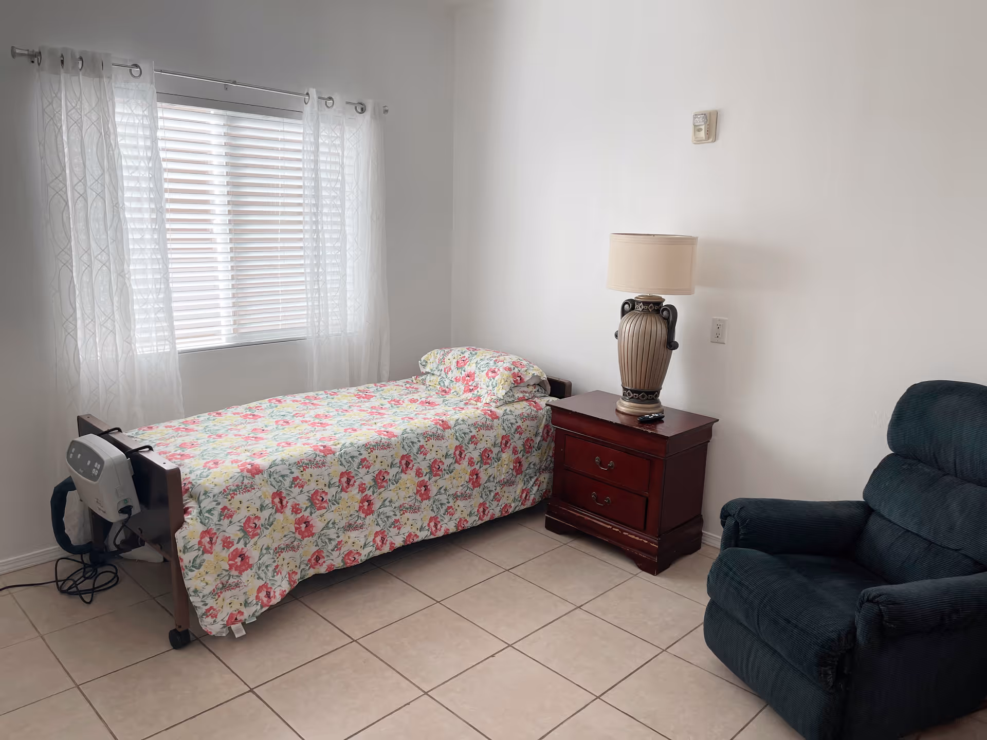 A simple bedroom with a single bed covered in a floral bedspread, a wooden nightstand with a decorative lamp, a dark green recliner chair, and a window with white curtains allowing natural light into the room.