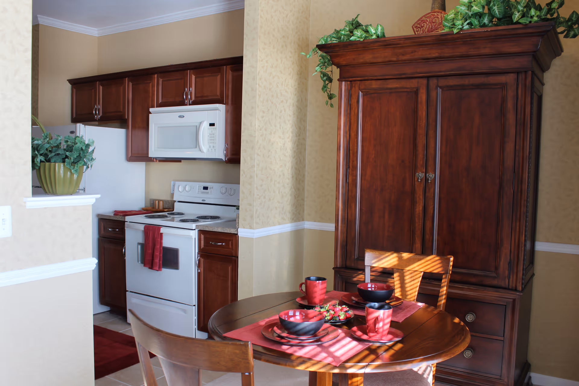 A cozy kitchen and dining area with wooden cabinets, a white stove and microwave, a round wooden dining table set with red and black dishes and cups, and a large wooden cabinet with green plants on top. Sunlight streams through blinds, casting shadows on the table and chairs.