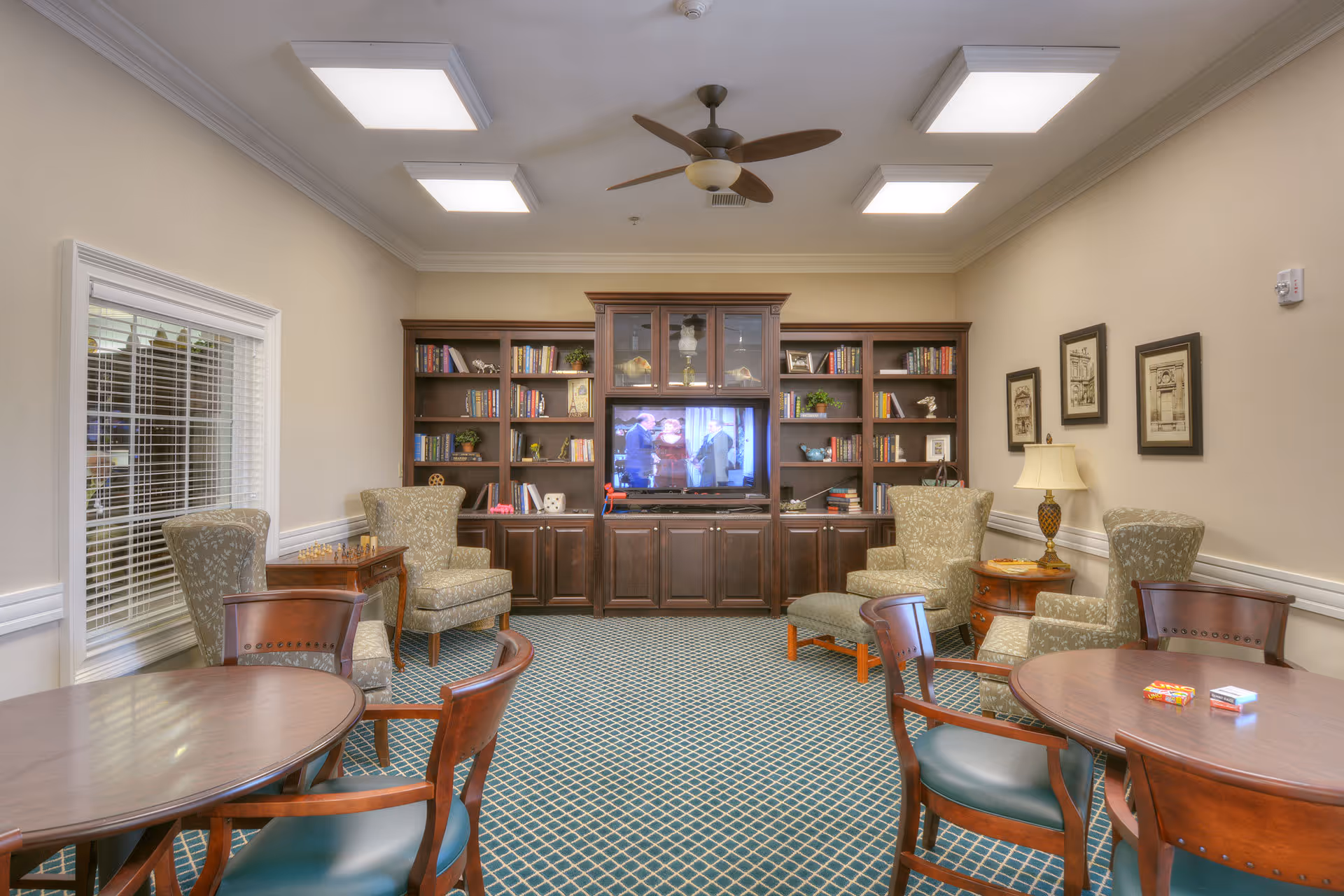 A cozy living room area in a senior living facility featuring a large wooden entertainment center with a TV, bookshelves filled with books and decorative items, several upholstered armchairs, round wooden tables with chairs, a ceiling fan, and framed artwork on the walls.