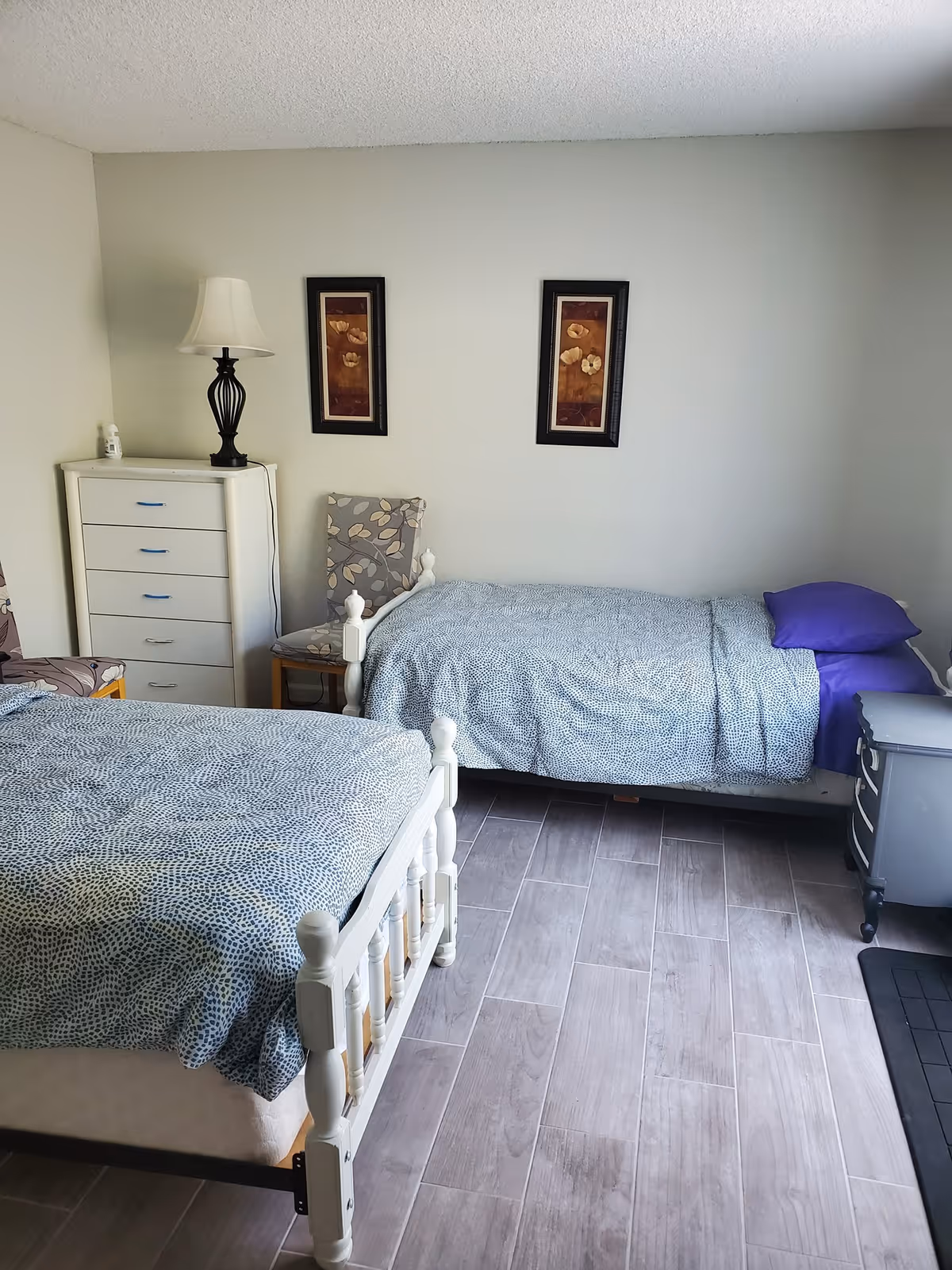 A simple bedroom with two single beds covered in blue patterned bedspreads. There is a white chest of drawers with a lamp and a bottle on top, a chair with a floral cushion, and two framed floral artworks hanging on the wall. The floor is covered with wood-like tiles.