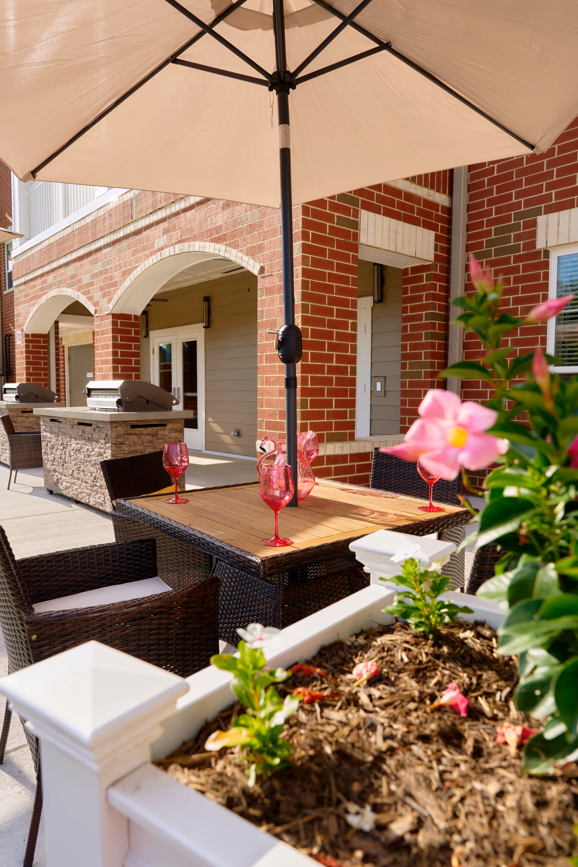 Outdoor patio area with a wooden table and four wicker chairs under a large beige umbrella. The table has four red wine glasses and two pink flamingo decorations. In the foreground, there is a white planter box with green plants and pink flowers. The background shows a brick building with arched doorways and two built-in grills.