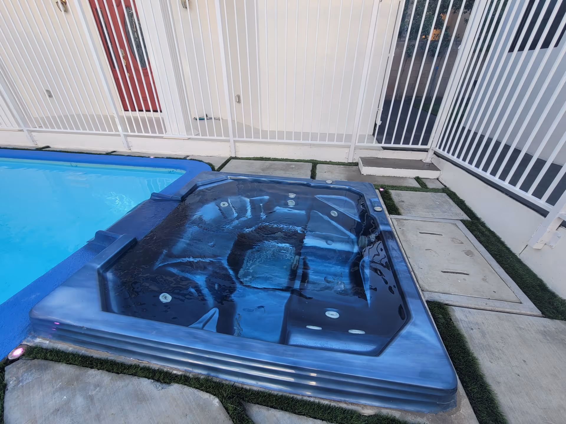Outdoor area featuring a blue swimming pool adjacent to a black hot tub with water inside, surrounded by a white metal fence and concrete tiles with patches of grass.