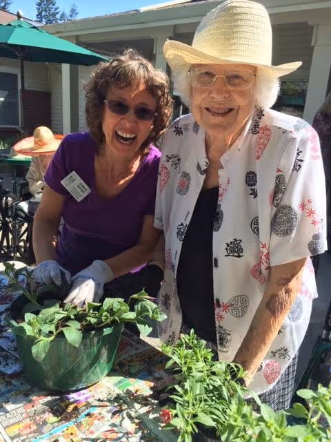 Two elderly women smiling and gardening outdoors at a table with green plants. One woman is wearing a white hat, glasses, and a patterned shirt, while the other woman is wearing sunglasses, gloves, and a purple shirt with a name tag. They are in a sunny outdoor area with a building and green umbrella in the background.