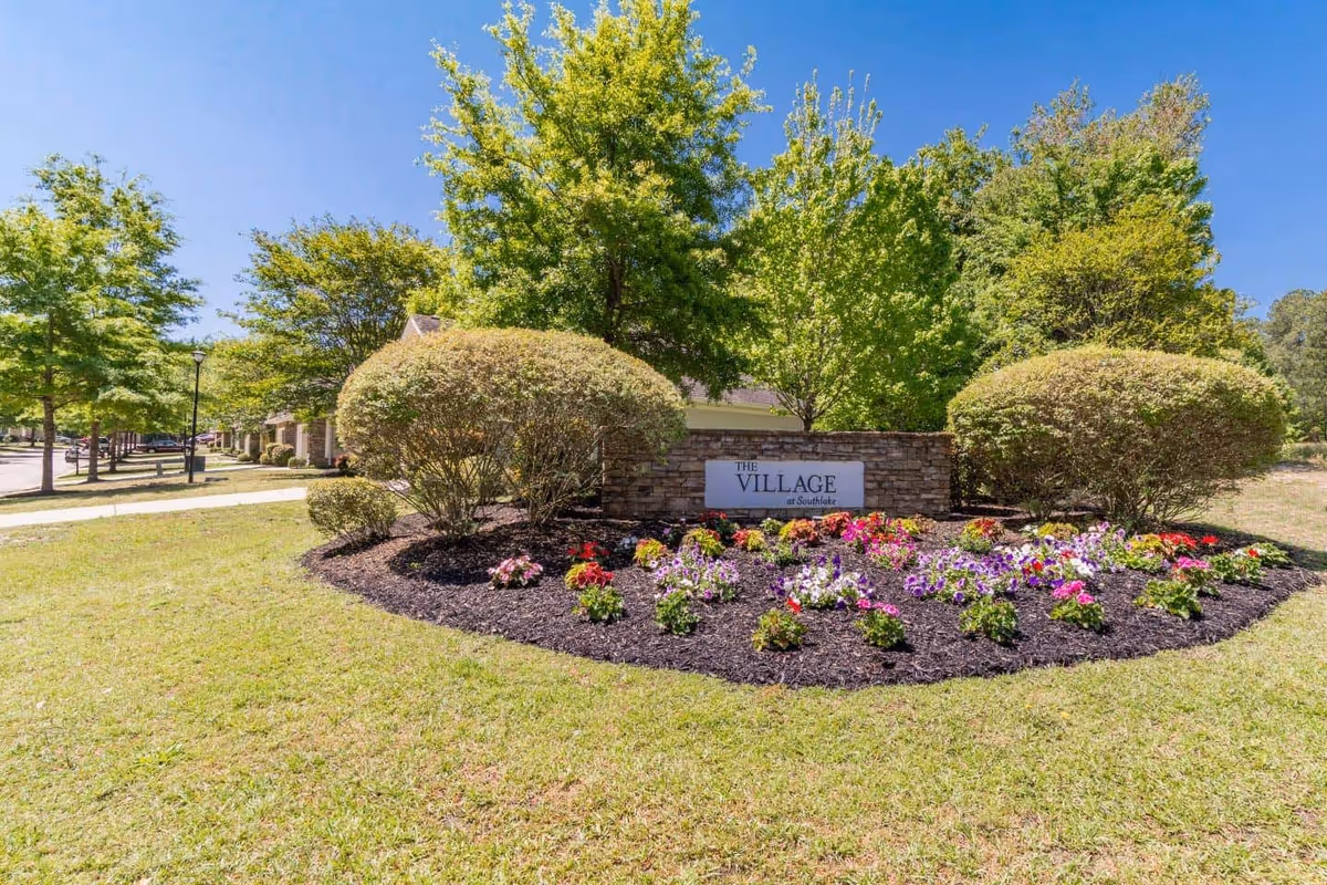 Landscaped entrance sign reading "The Village at Southlake" surrounded by flowers, shrubs, and trees under a blue sky.