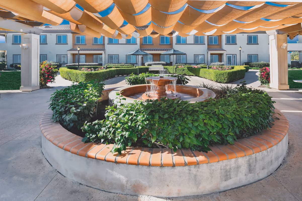 A shaded courtyard with a central circular fountain surrounded by plants and hedges in front of a multi-story building.