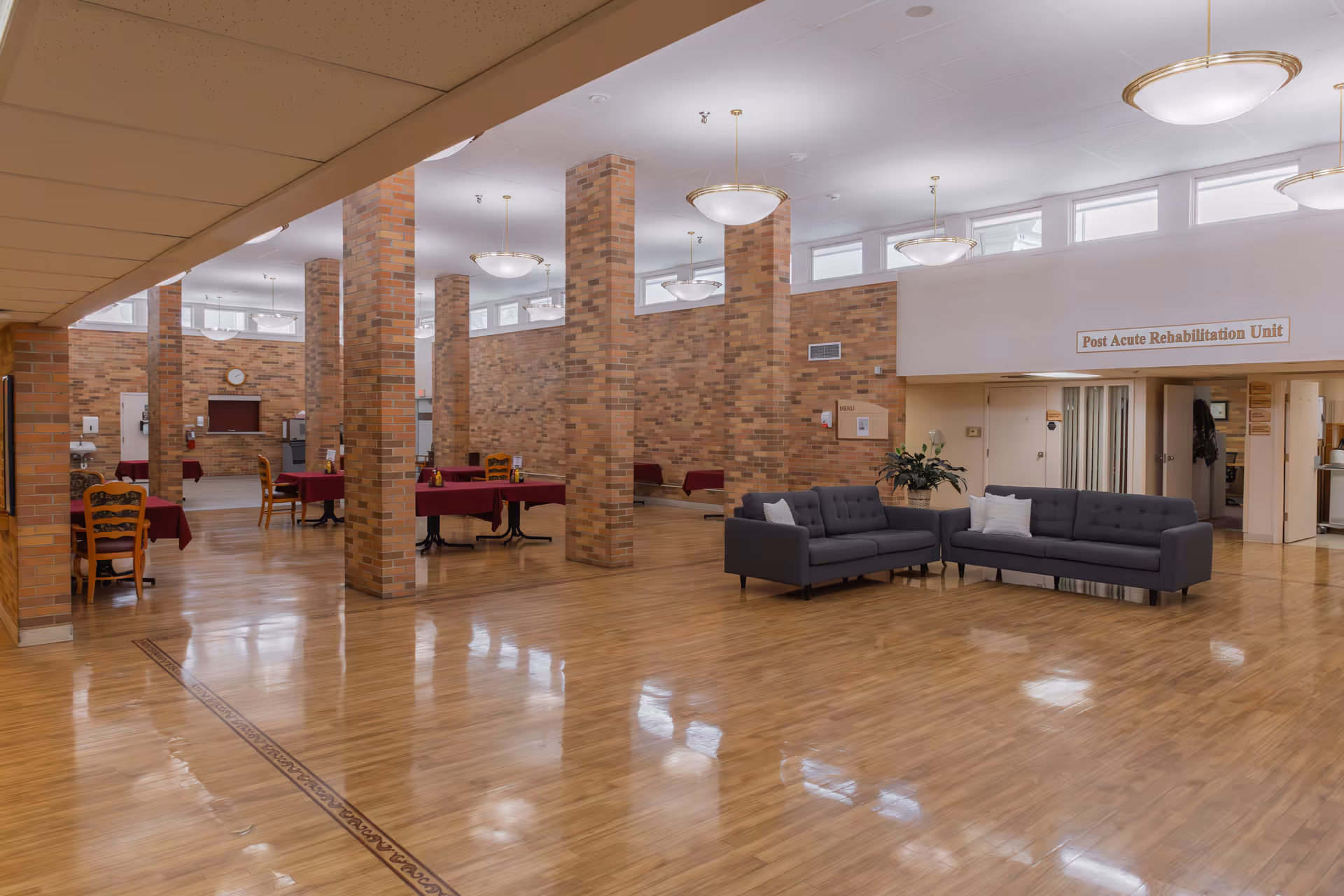 Interior view of a senior living facility with polished wooden floors, brick pillars, and several tables with red tablecloths. There are two gray sofas with a small table and plant between them. A sign on the wall reads 'Post Acute Rehabilitation Unit'. The ceiling has multiple round light fixtures and high windows near the ceiling.