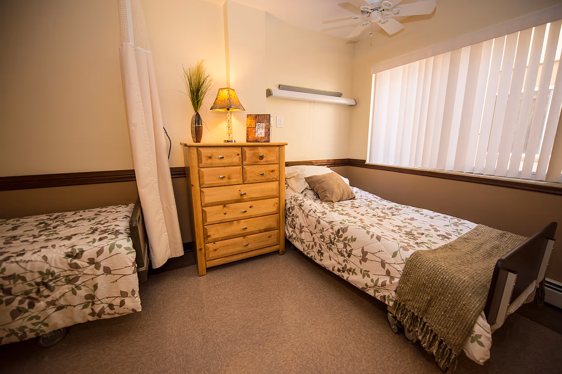 A small bedroom in a care facility with two single beds separated by a wooden chest of drawers. Each bed has floral patterned bedding and pillows. A lamp, a framed picture, and a vase with tall grass are on top of the chest of drawers. The room has beige walls, a window with vertical blinds, and a ceiling fan.