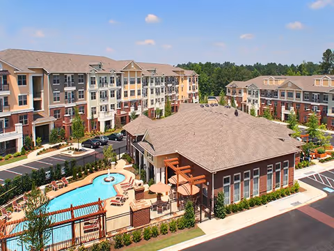 Aerial view of a senior living facility named Park at Johns Creek featuring multi-story residential buildings surrounding a central building with a fenced swimming pool and outdoor seating area. The scene includes landscaped greenery, parking spaces, and a clear blue sky.