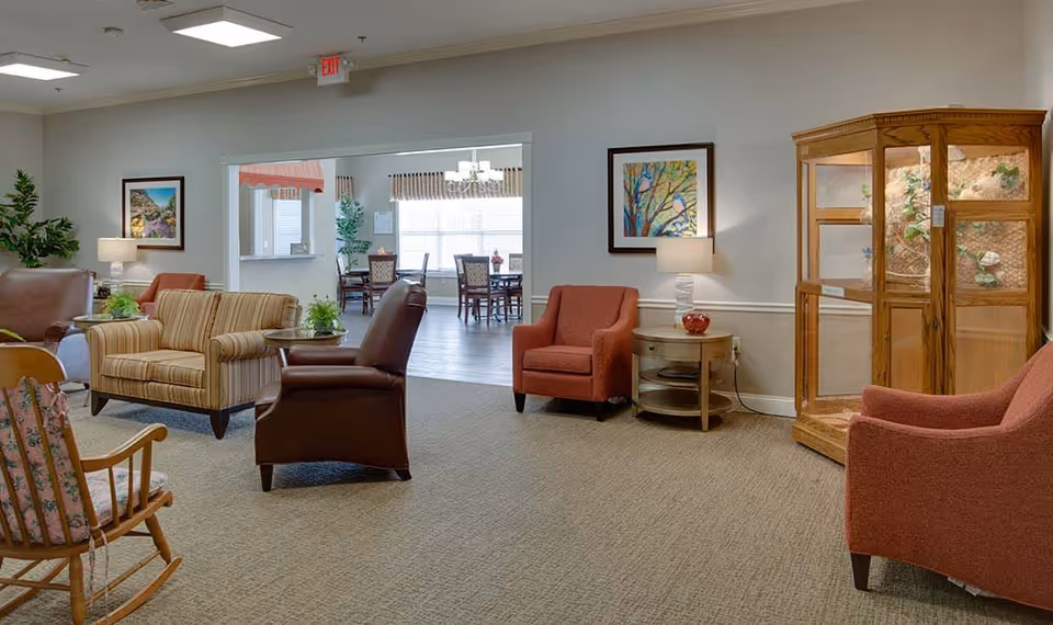 A cozy senior living facility common area with various seating options including a striped loveseat, brown leather chair, and two red armchairs. There is a wooden rocking chair in the foreground and a wooden display cabinet on the right. The room has beige carpeting, light gray walls with framed artwork, and a side table with a lamp. In the background, a dining area with tables and chairs is visible through an open doorway.