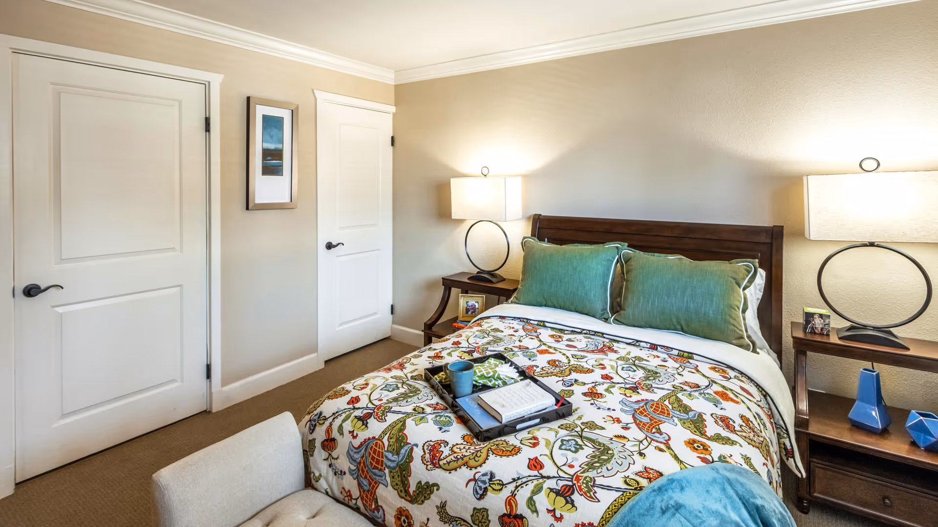 A cozy bedroom with a double bed featuring a colorful floral patterned bedspread and two green pillows. There are two matching wooden nightstands on either side of the bed, each with a modern lamp. The room has beige walls, two white doors, and a small framed picture hanging between the doors. A beige armchair is partially visible in the foreground.