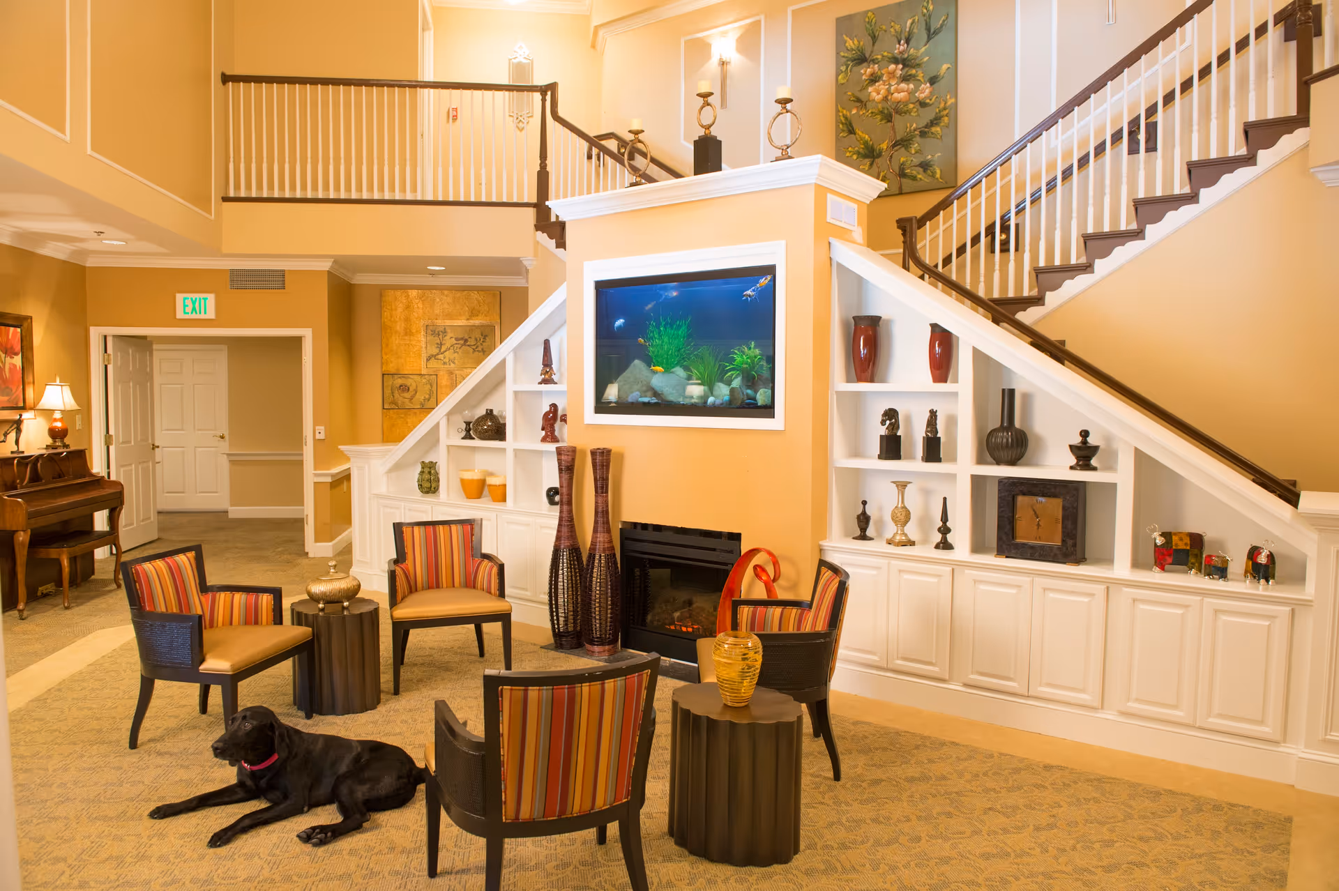 A cozy living room area in a senior living facility with four striped armchairs arranged around two small tables. A black dog is lying on the carpeted floor. The room features built-in white shelves under a staircase, decorated with various vases, sculptures, and a clock. There is a fireplace with an aquarium above it. The walls are painted a warm beige color, and there is a piano and lamp in the background near an exit door.