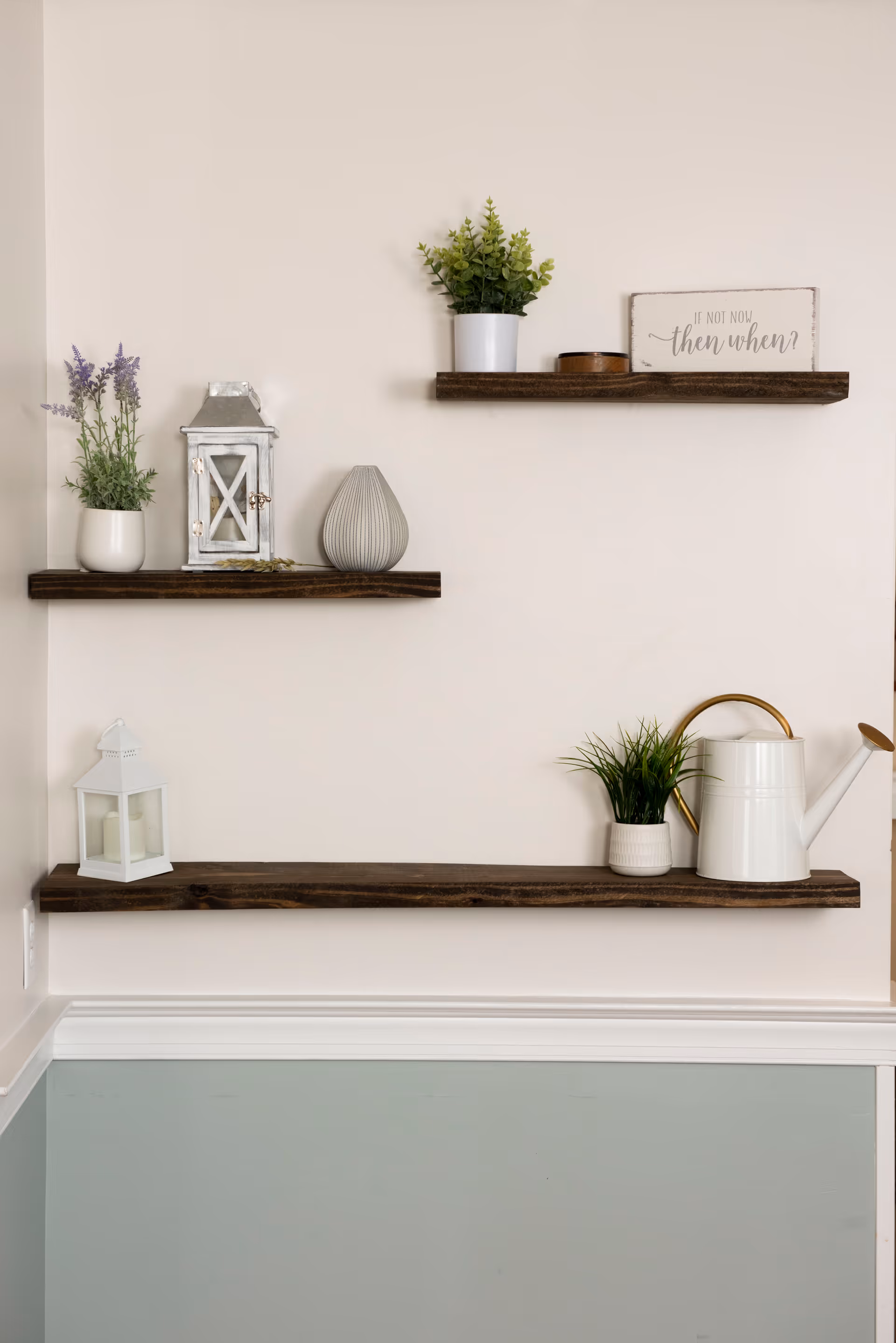 Three dark wooden floating shelves mounted on a beige wall above a light blue wainscoting. The shelves display decorative items including potted plants, a white lantern, a small watering can, a gray vase, and a sign that reads 'If not now then when?'.